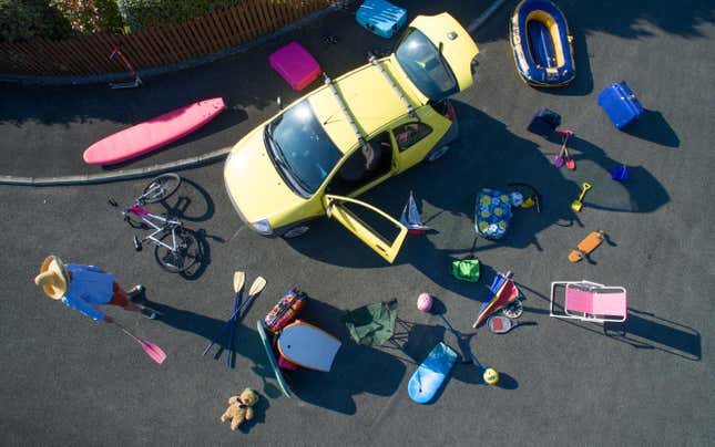 An overhead view of a yellow car parked on blacktop with an array of vacation stuff spread out around it.