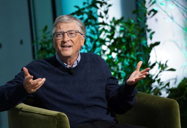 Bill Gates sitting in a chair with both his arms up as if he's confused