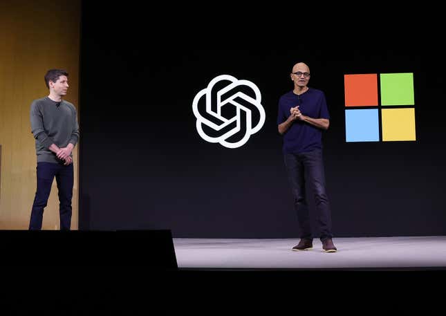 Sam Altman wearing a grey sweater and black pants standing near Satya Nadella wearing a navy top and jeans in front of a backdrop with the OpenAI and Microsoft logos