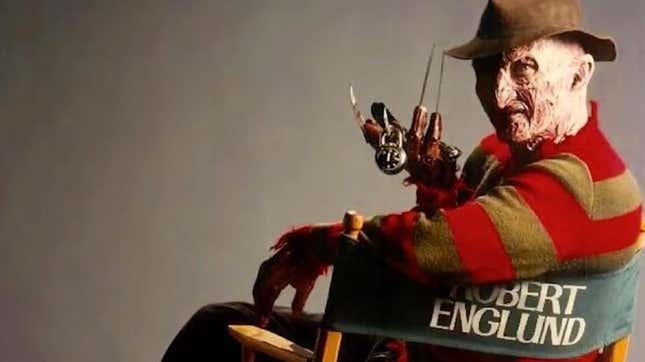 Robert Englund sits in a director's chair with his name on it, dressed in full Freddy Krueger make-up and costume.