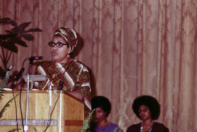 Audre Lorde talking on microphone and Alice Walker and June Jordan sitting in background at The Phillis Wheatley Poetry Festival.