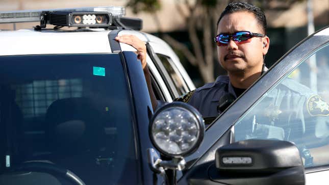 A male police officer wearing sunglasses is stepping out of his patrol car, his hand on the roof near an automatic license plate reader mounted to the cruiser.