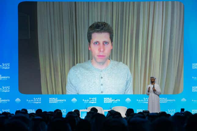 Sam Altman wearing a grey shirt with buttons on screen surrounded by blue while a man on stage in front of him speaks to the crowd