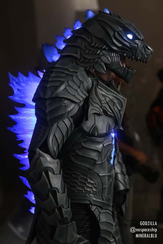 A cosplayer dons a full Godzilla suit, complete with glowing blue spikes on his back. 