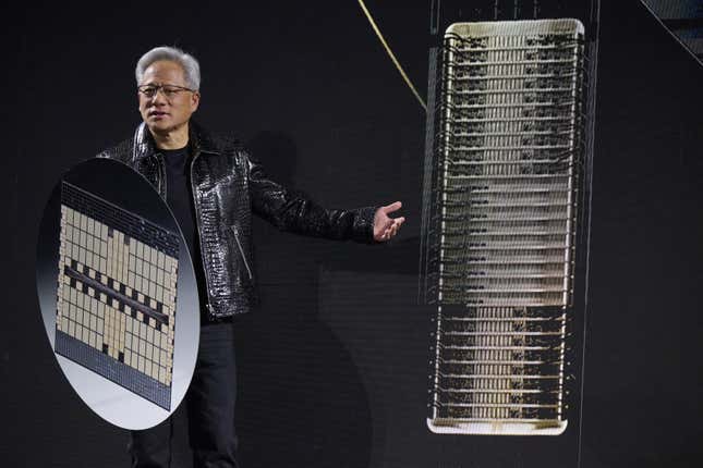 Jensen Huang wearing a black t shirt and black leather jacket standing in front of a backdrop displaying a rack and holding a large circle with an enlarged chip on it