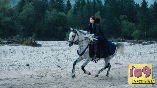 Rosamund Pike, as the magic user Moraine, rides a white horse with black splotches past a forest in a scene from Wheel of Time.