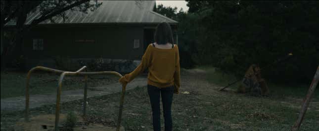 A woman facing away from the camera looks toward a roadside rest stop building that appears to be abandoned in horror short Seek.