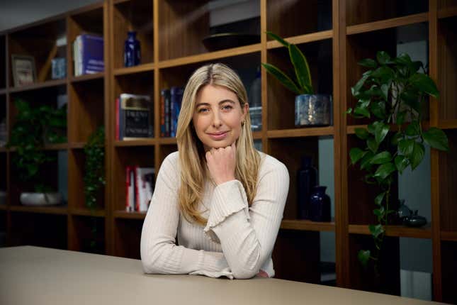 a blonde woman sitting and smiling at the camera wearing a white shirt, her left fist is under her chin