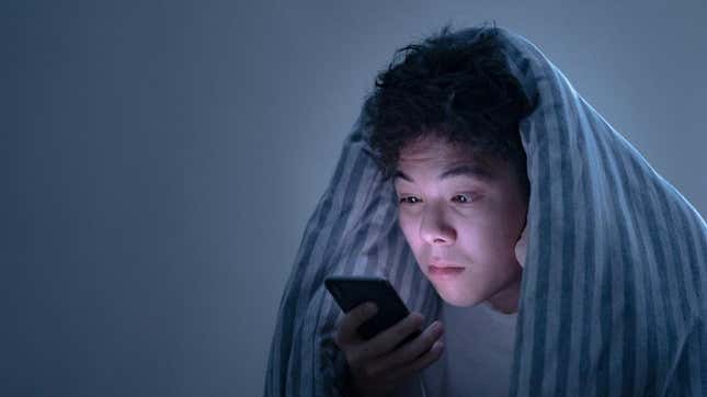 Ein müder Mann unter einer Decke, der auf ein Telefon starrt