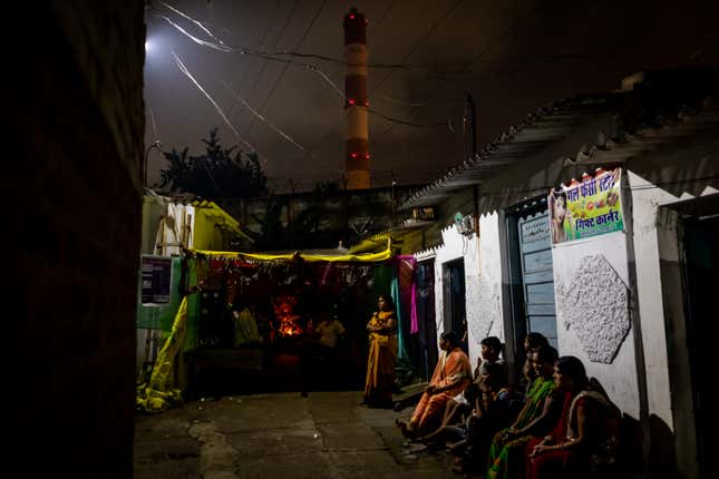 Residents of Patharripara neighborhood in Korba wait to resume religious festivities during a power outage, as the chimney of a coal power plant blows out smoke in the background. They were still waiting when we left the area half an hour later.