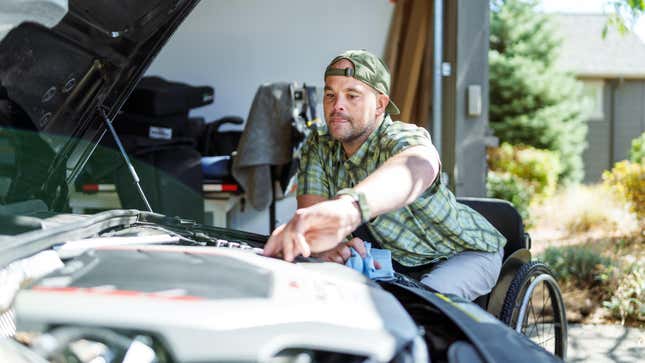 Un homme d’âge moyen handicapé qui utilise un fauteuil roulant travaille sur sa voiture à la maison, dans le garage, par une journée ensoleillée.