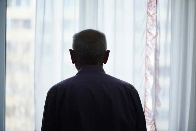 Silhouette of retired man looking through window with transparent curtain standing at home rear view. 