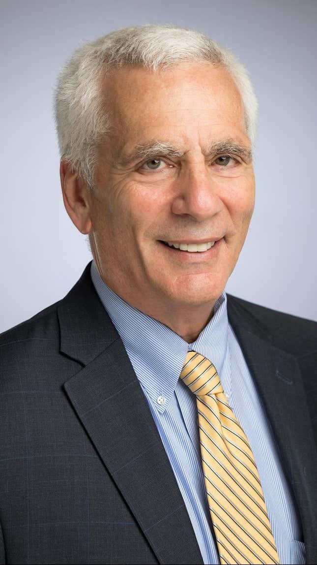 Jared Bernstein stands smiling in front of a grey background wearing a dark grey jacket, yellow striped tie, and light blue shirt.