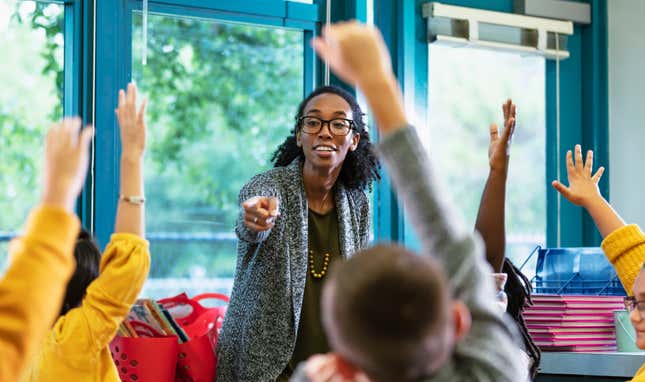 A multiracial group of elementary students in class raising their hands to answer a question. T