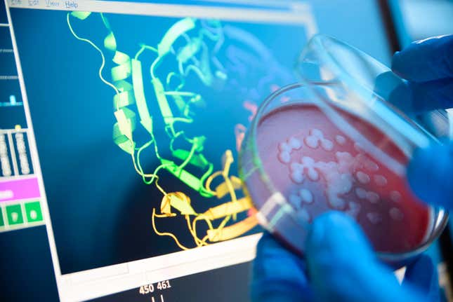 gloved hands holding a petri dish in front of a monitor displaying structural models of proteins