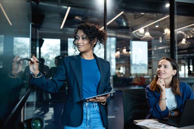 a woman sitting looks on as another woman presents market data on a digital display