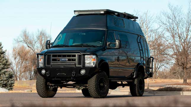 A Custom Ford Camper Van in black parked in a parking lot surrounded by winter trees.