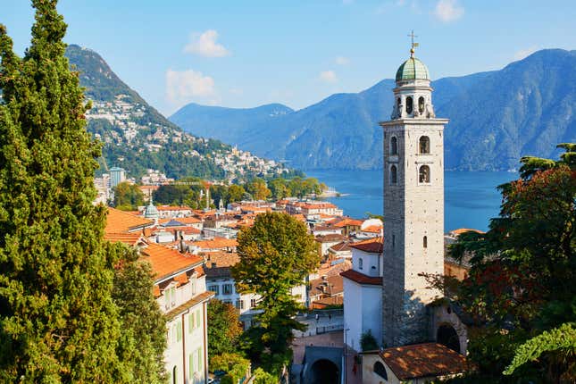 scenic view of old town Lugano with a church bell tower in front of a body of water and mountains