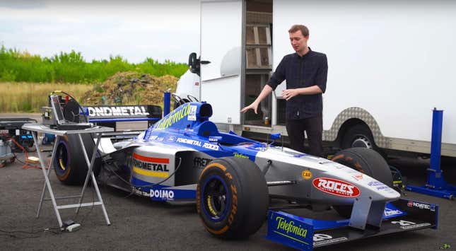 A 1998 Formula 1 car is parked near a race hauler preparing to be started. A slim man is pointing at it.