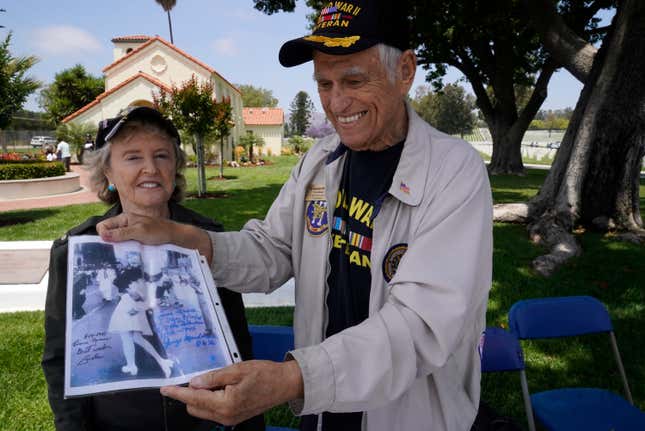 World War II U.S. Army veteran Dr. George Jack Stanley, with his wife,  Sandra Stanley, members of The Los Angeles National Cemetery Support  Foundation, welcome veterans to the Los Angeles National Cemetery, in  Los Angeles, Monday, May 31, 2021. Stanley is displaying a copy of the  famous “V-J Day in Times Square,” a photograph by Alfred Eisenstaedt,  autographed by the picture’s subjects: Greta Zimmer Friedman and George  Mendonsa. 