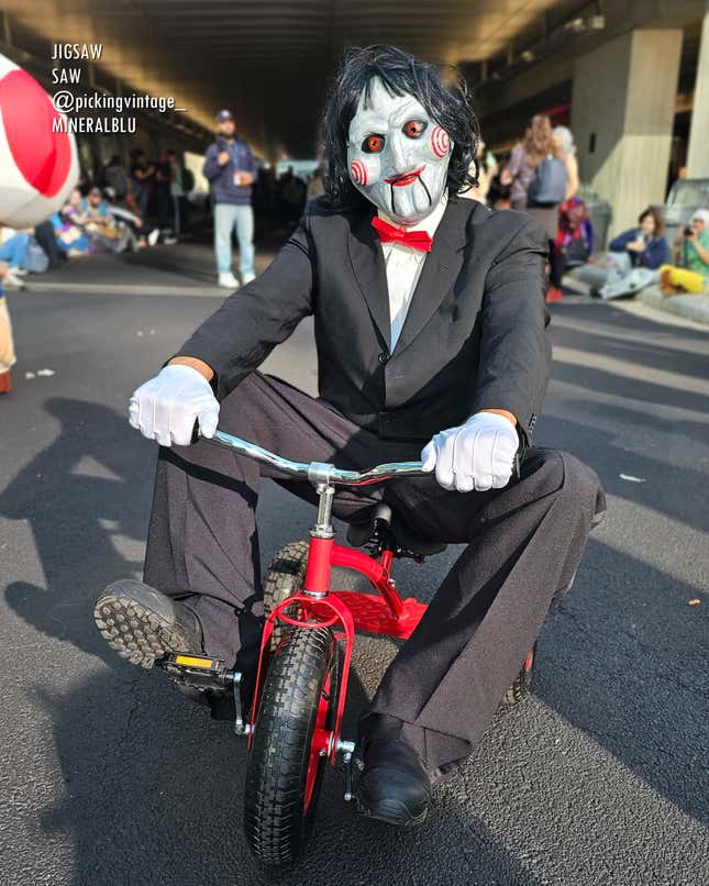 A person dressed as Jigsaw from the Saw movies rides a kid's tricycle. 