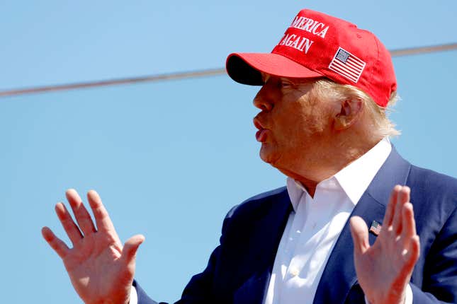 Donald Trump, in a red hat and with both palms facing out, speaks