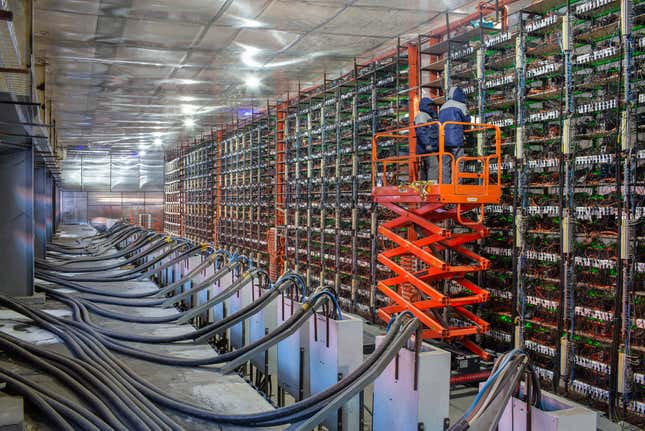 engineers on an orange cherry picker inspecting a row of mining rigs