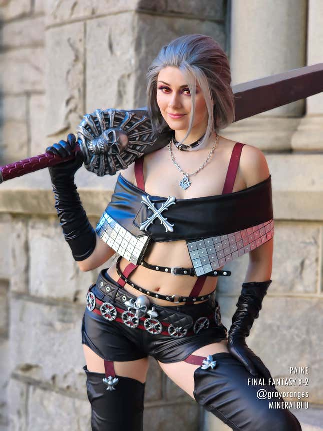 A cosplayer dressed as Paine from Final Fantasy X-2 stands with her sword over her shoulder and a smirk on her face.
