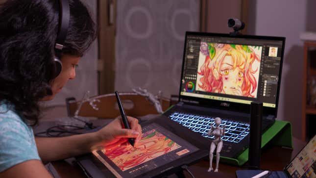 A young woman wearing headphones uses a stylus on an art program on a tablet while a laptop in front of her shows the same image.