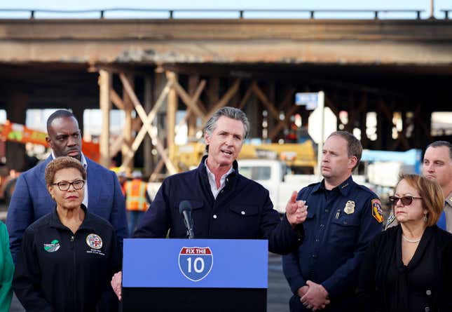 Governor Gavin Newsom speaks in front of the charred freeway overpass as Los Angeles Mayor Karen Bass listens at a press conference near the closed I-10 elevated freeway following a large pallet fire, which occurred Saturday at a storage yard beneath the freeway, on November 13, 2023 in Los Angeles, California.