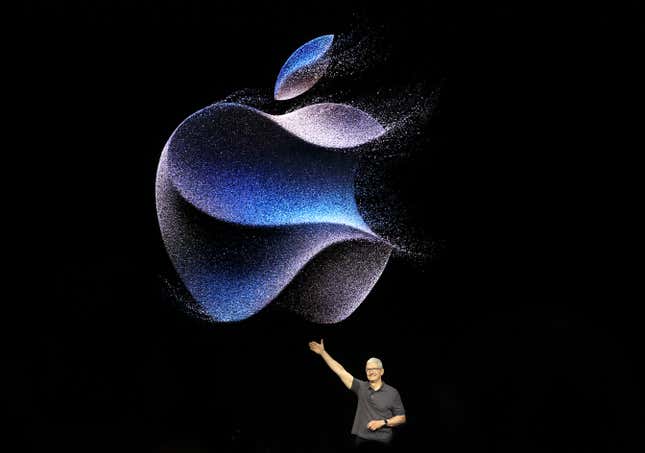 Tim Cook is small standing in the center-right of the photo showing him on stage in front of a large apple logo on a backdrop