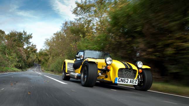 A photo of a yellow Caterham driving on a country road. 