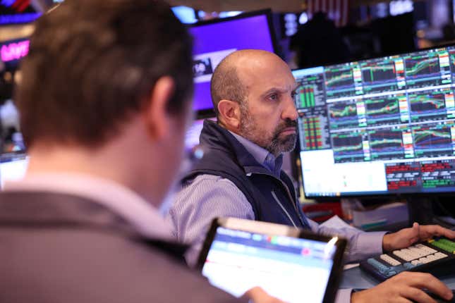 Two men, one with his back to the camera, look at trading figures on computer screens