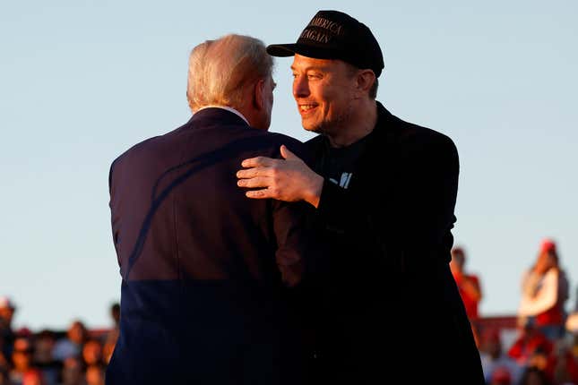 Donald Trump's back is facing the camera as he hugs Elon Musk whose face can be seen, blurry people are in the background