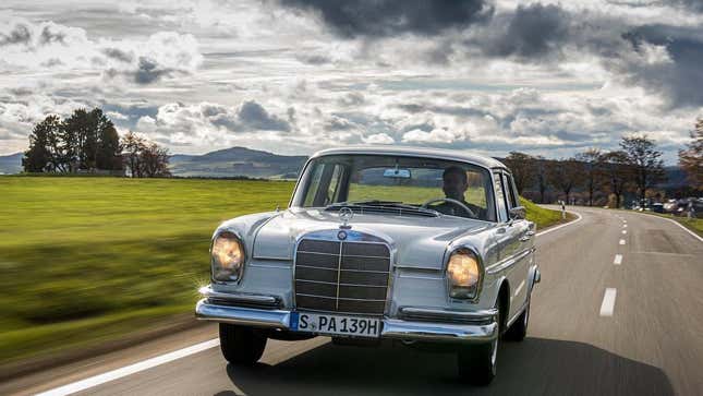 A 1961 Mercedes 300 SE driving in front of a beautiful cloudy sky and a green grassy field