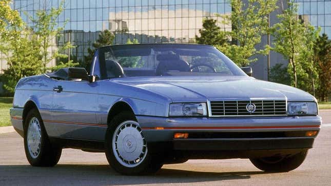 A silver Cadillac Allante with its top down in front of a very 80s looking office building