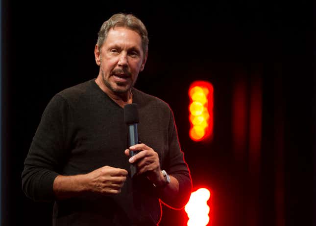 Larry Ellison wearing a black long sleeved shirt holding a microphone and speaking into it in front of a black backdrop with reddish lights in the background