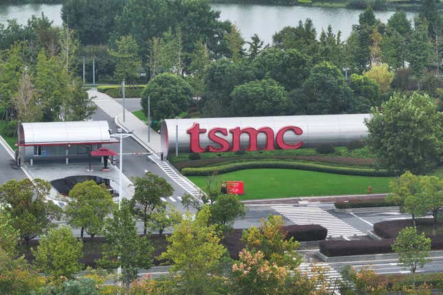 aerial view of part of the TSMC factory surrounded by green trees and a lake in the background, red tsmc letters appear on the side of a metal tube