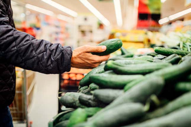 Hombre de pie frente a un puesto de productos agrícolas y eligiendo pepinos