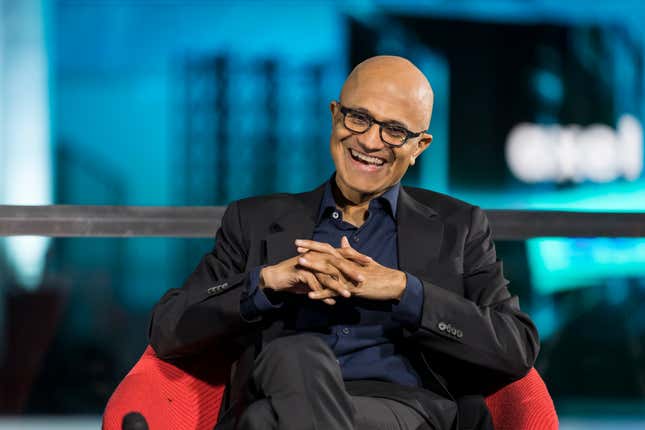 Satya Nadella wearing a black suit jacket, black dress pants, and navy button down, sitting in a red chair laughing with his hands clasped together and waring glasses