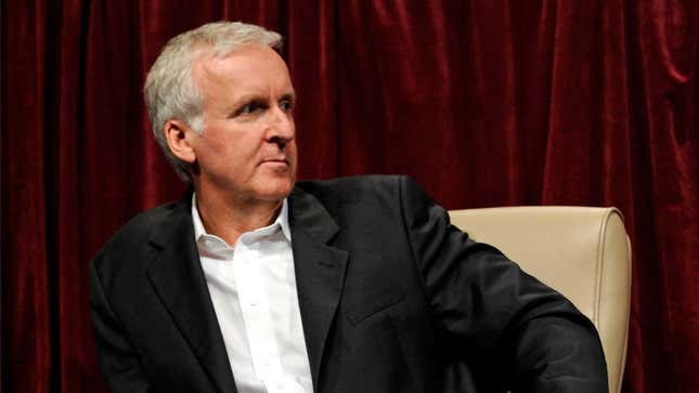 James Cameron, in a white-button-down shirt and black jacket, sits in front of red movie curtains.