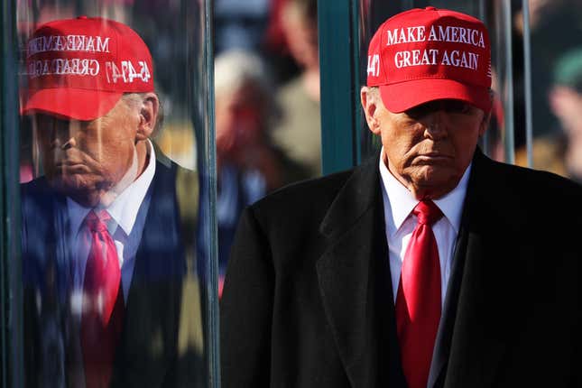 Donald Trump lleva una gorra de Make America Great Again, lleva traje y corbata, su rostro está parcialmente oculto por una sombra y se refleja en un vidrio a su lado.