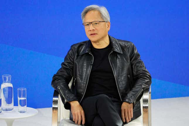 Jensen Huang wearing all black sitting in a chair in front of a royal blue backdrop