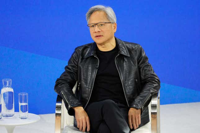 Jensen Huang vestido todo de negro sentado en una silla frente a un fondo azul real