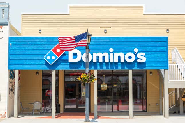 Outside of a Domino's location — cream and blue slatted wood with a small seating area out front and the Domino's logo