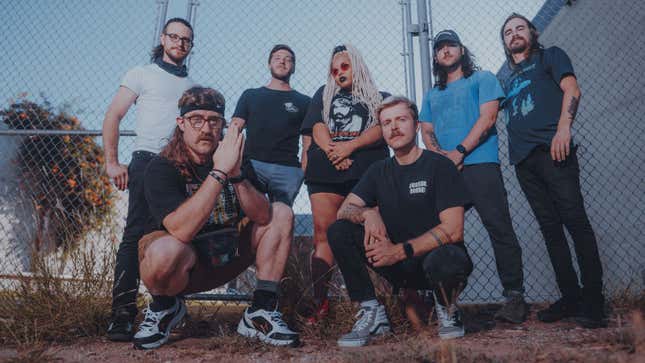 A seven-person band poses in front of a chain link fence. There are many mustaches.