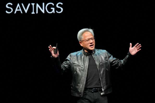 Jensen Huang wearing all black including a leather jacket speaking in front of a black backdrop that has SAVINGS written in white at the top left corner