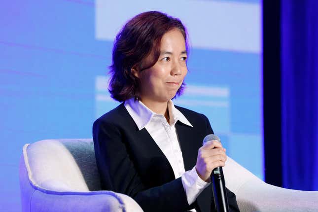 Fei-Fei Li wearing a white button down and black jacket sitting in a grey chair holding a microphone, the backdrop behind her is purple