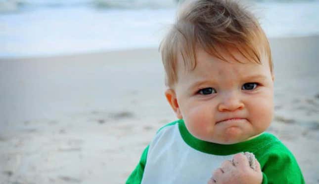 A white baby in a green and white baseball t-shirt makes a fist on a beach.