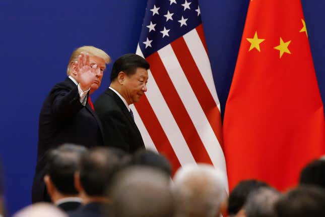 Trump saluda con la mano, ocultándose parcialmente el rostro, y camina un poco detrás de Xi Jinping, cuyo perfil derecho se puede ver. Ambos caminan frente a banderas estadounidenses y chinas, una al lado de la otra.